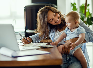 Woman Holding baby while siting at desk with a pen and a laptop