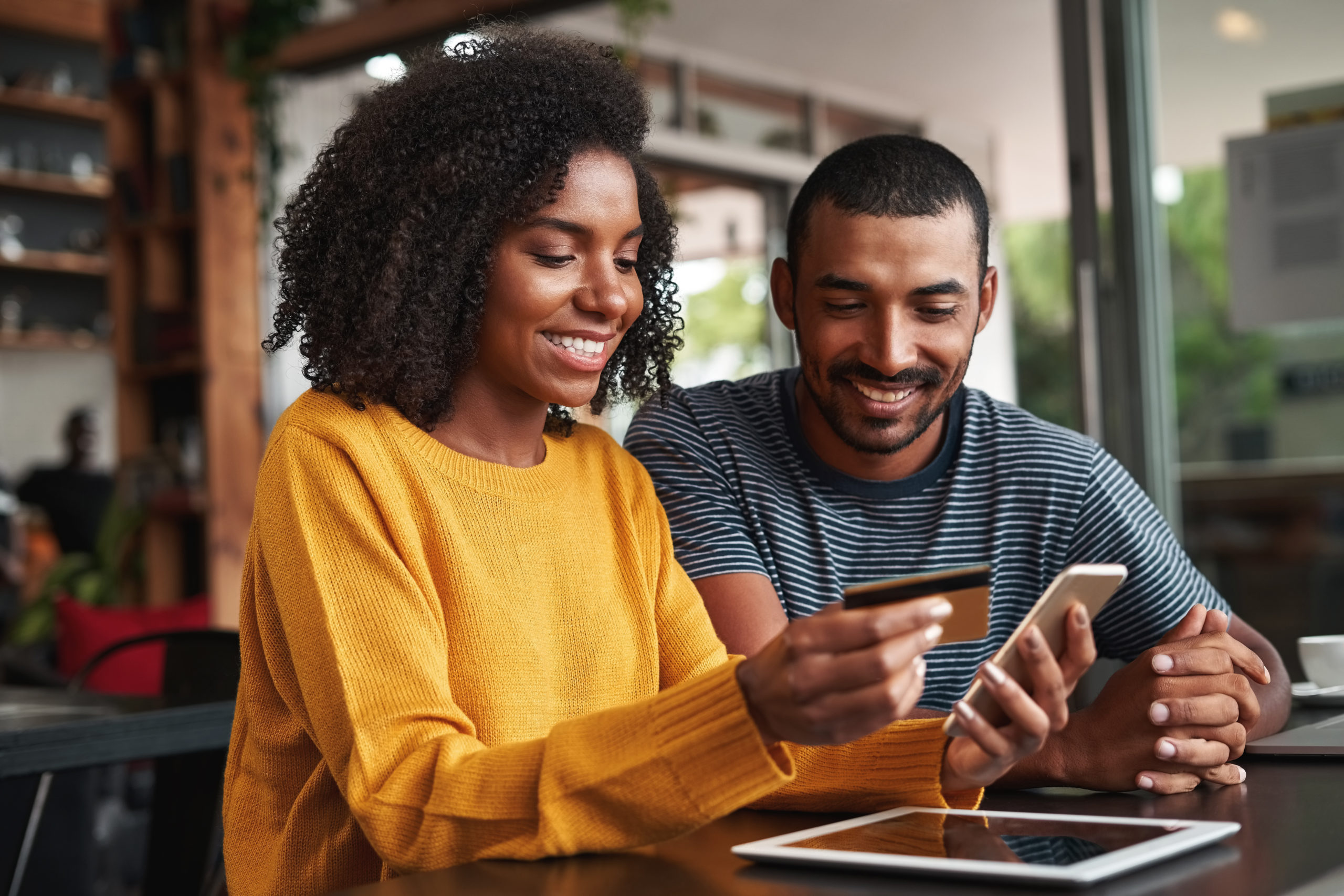 Young african man looking at his girlfriend using credit card and smartphone for shopping online in cafe