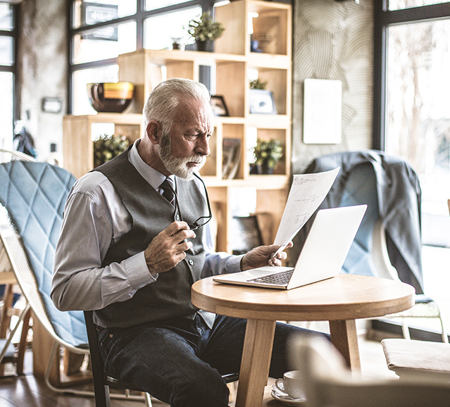 man siting at a small round desk with a laptop, paper and reading glasses