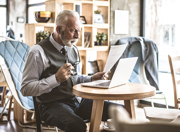 man siting at a small round desk with a laptop, paper and reading glasses